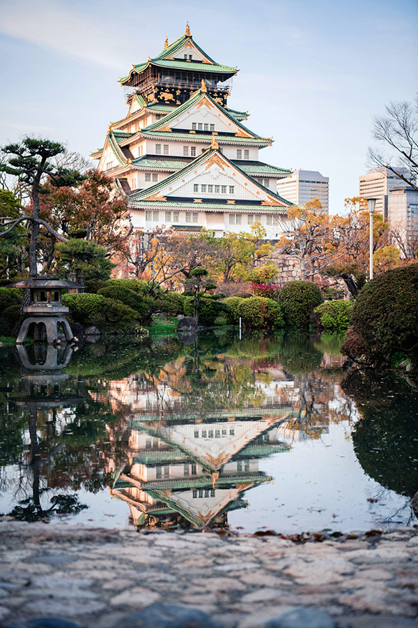 Osaka Castle reflected in a garden pond, surrounded by autumn trees and traditional Japanese landscaping.
